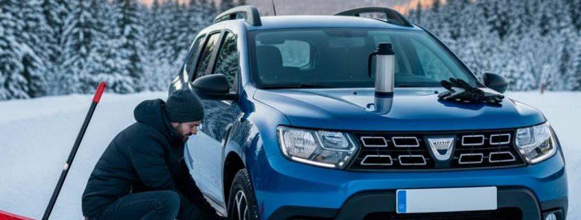 Hombre colocando cadenas de nieve en un Dacia Duster azul en una carretera de montaña nevada, con una pala roja y un termo al lado.